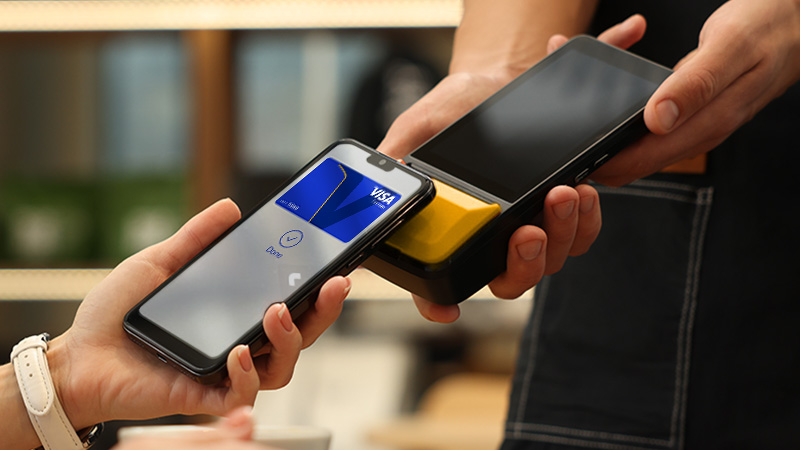 A person completes a contactless payment using a smartphone with a Visa card displayed on the screen, tapping a card reader held by a cashier.