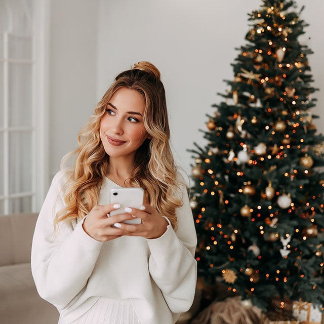 Woman with long blonde hair, wearing a white sweater, holds a smartphone and smiles. She stands by a decorated Christmas tree, creating a festive atmosphere.