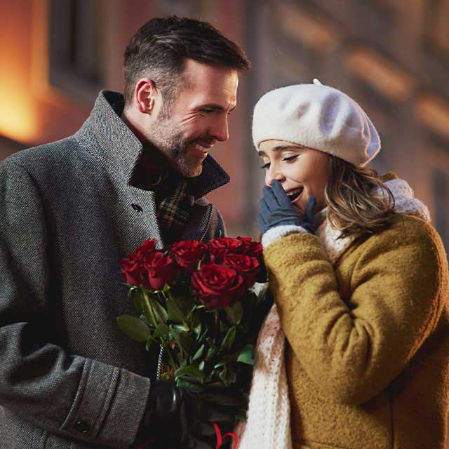 A man in a coat gives a bouquet of red roses to a delighted woman in a beret and scarf. They are outdoors, conveying warmth, romance, and surprise.