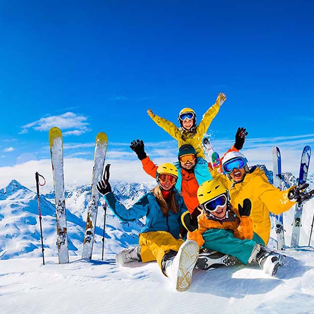 The company on a snowy mountain top smile at the camera, their skis are streaked in the snow nearby