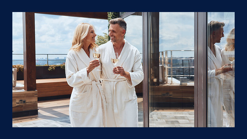 A couple in white bathrobes smiles at each other while holding wine glasses on a sunny rooftop, conveying relaxation and happiness. Reflections are visible in glass.