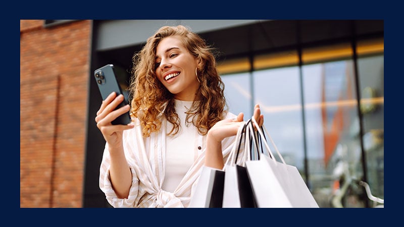 A smiling woman stands outside holding shopping bags and a smartphone, conveying excitement. She appears to be enjoying a successful shopping trip.